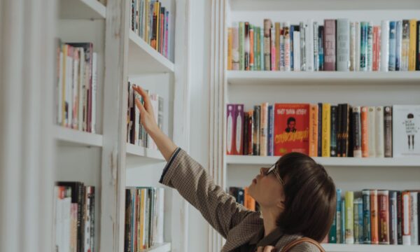 Woman in Beige Coat Standing in Front of White Wooden Book Shelf